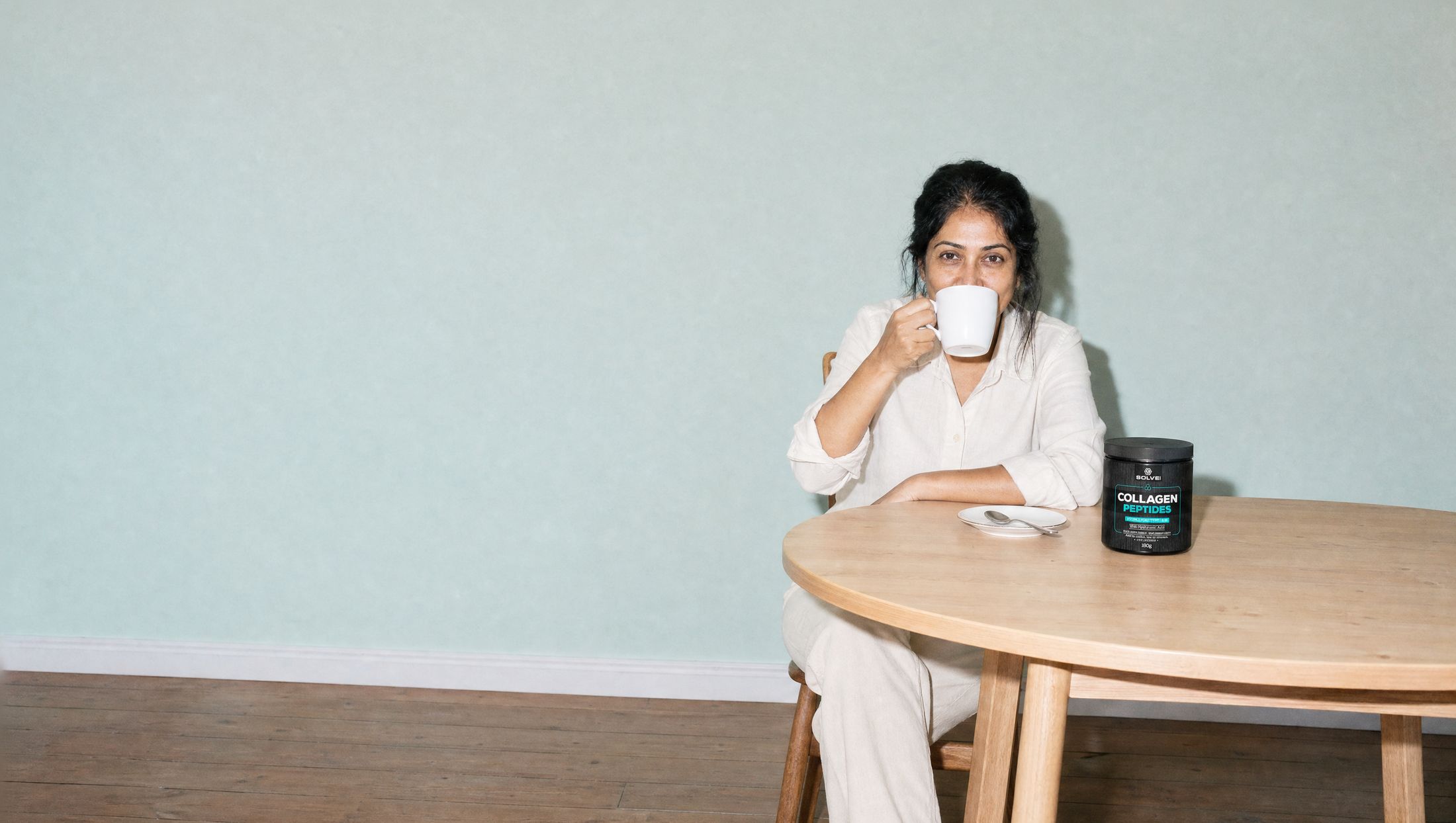 A woman at a kitchen table in the morning drinking a mug of coffee, the tub of Solve Labs Collagen Peptides beside her