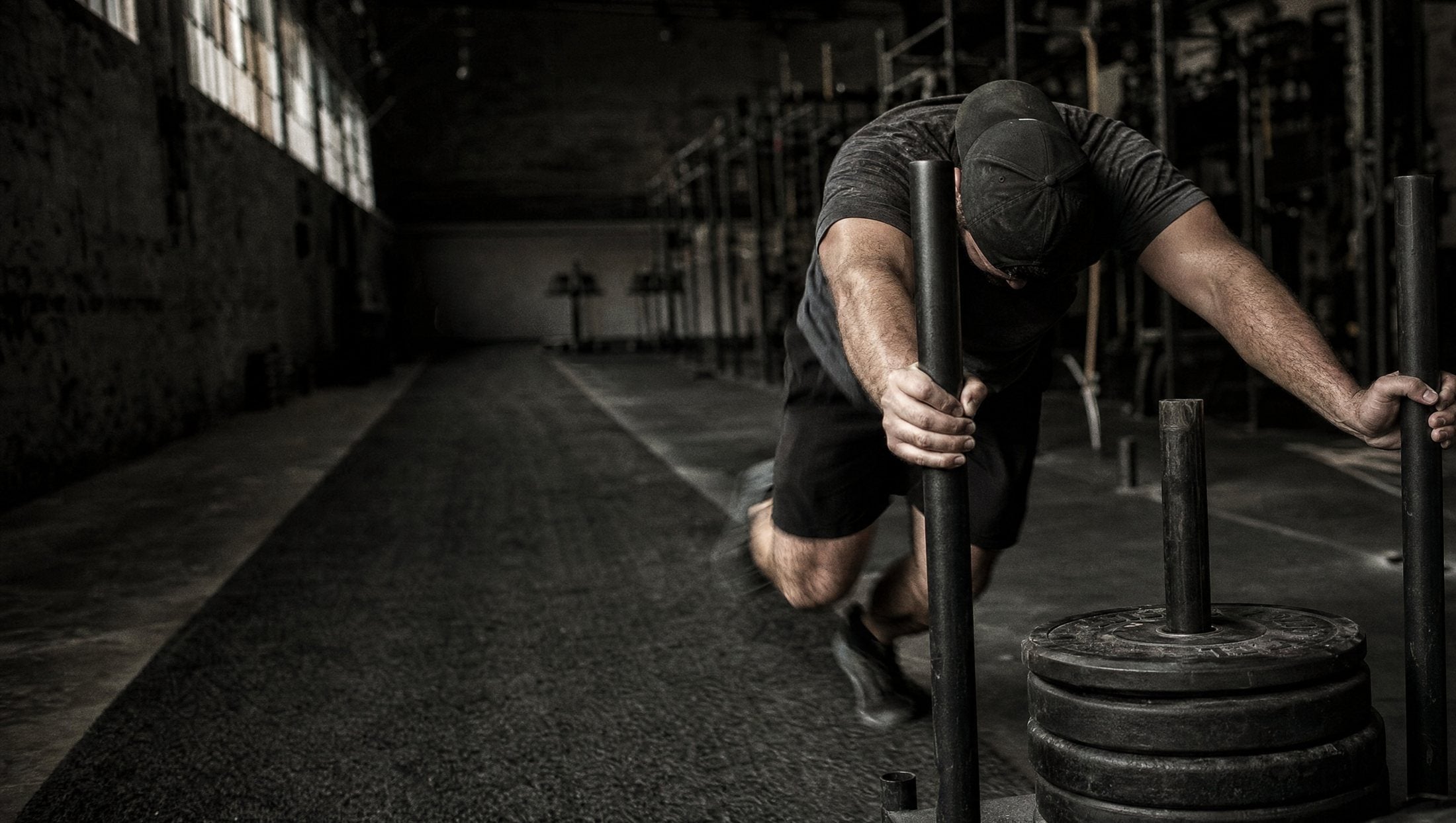 A man in a backwards cap driving a loaded push sled down the turf of a dark warehouse gym, head down