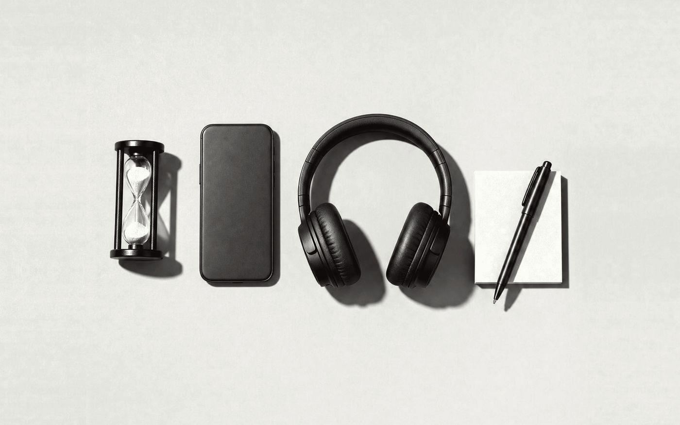 An hourglass, a phone face down, headphones and a notepad with a pen laid out in a row, in black and white