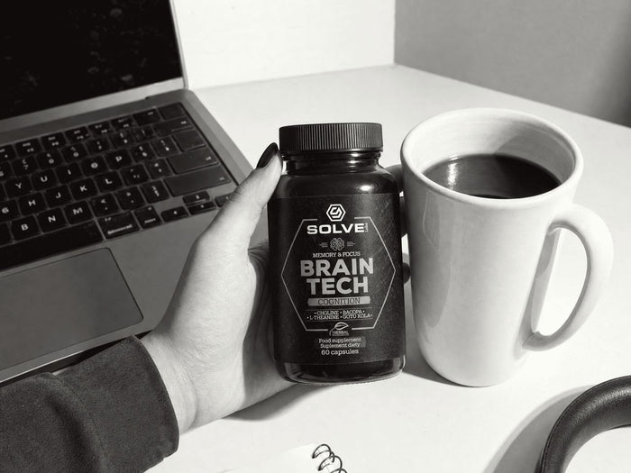 Eine Hand hält eine Dose Brain Tech neben einem Laptop und einem Becher Kaffee