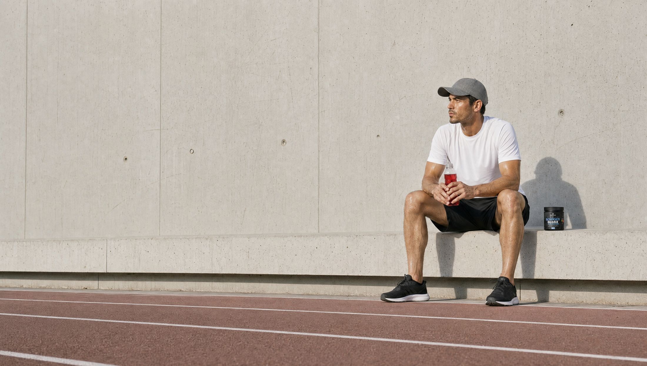 A runner sitting on a concrete bench beside an athletics track with a bottle of red drink, the tub of Solve Labs Electrolyte Balance beside him