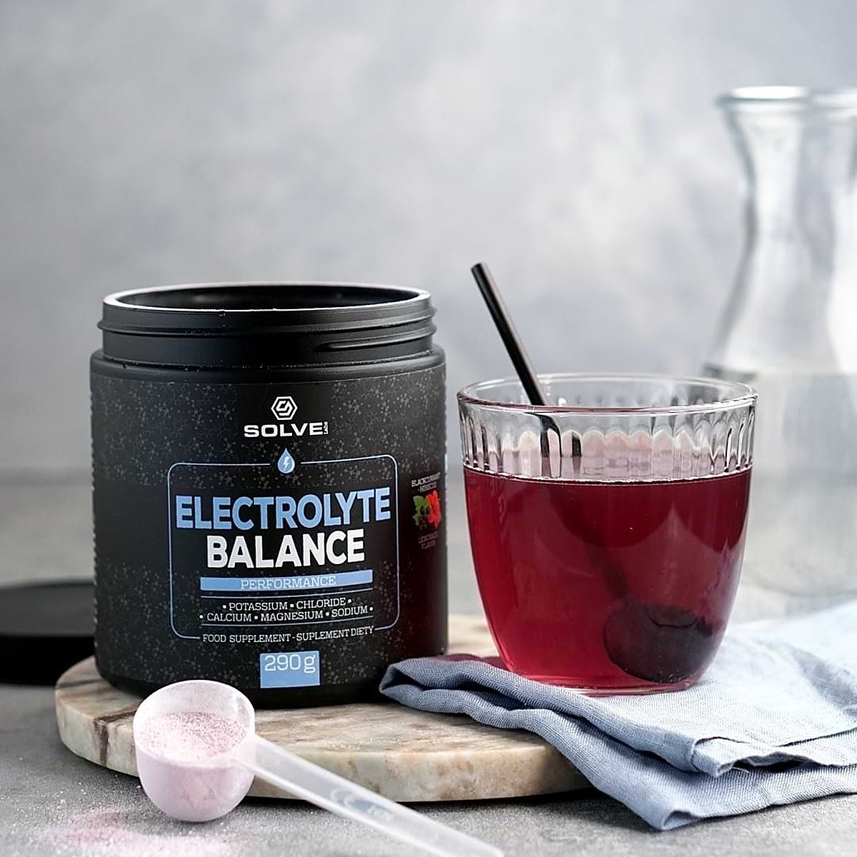 The tub of Solve Labs Electrolyte Balance on a marble board with a pink scoop of powder and a glass of the red drink