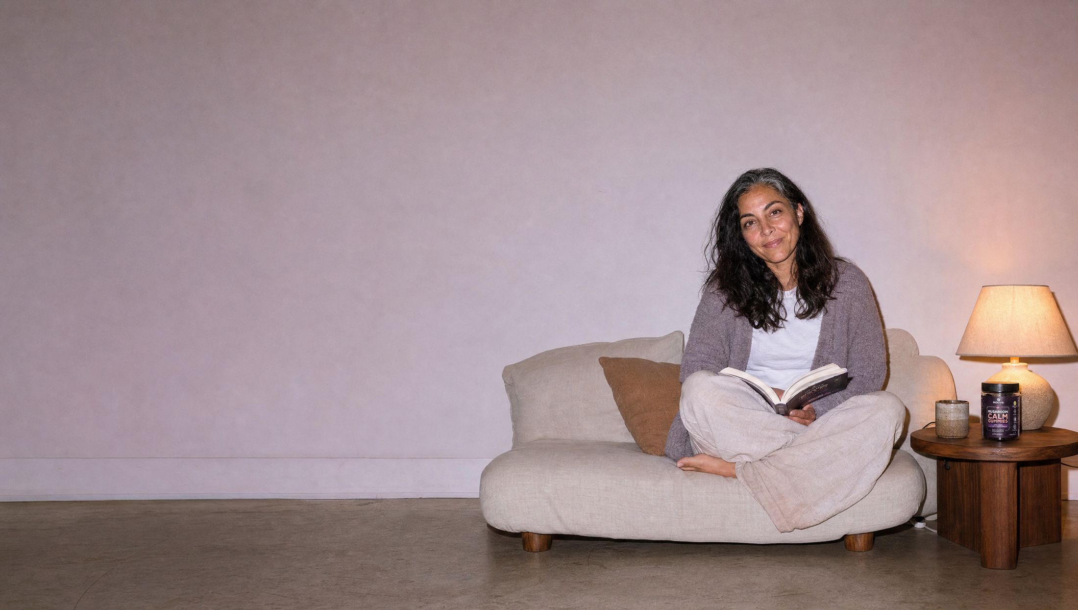 A woman curled on a sofa in the evening with a book, the jar of Solve Labs Mushroom Calm Gummies on the side table under a lamp