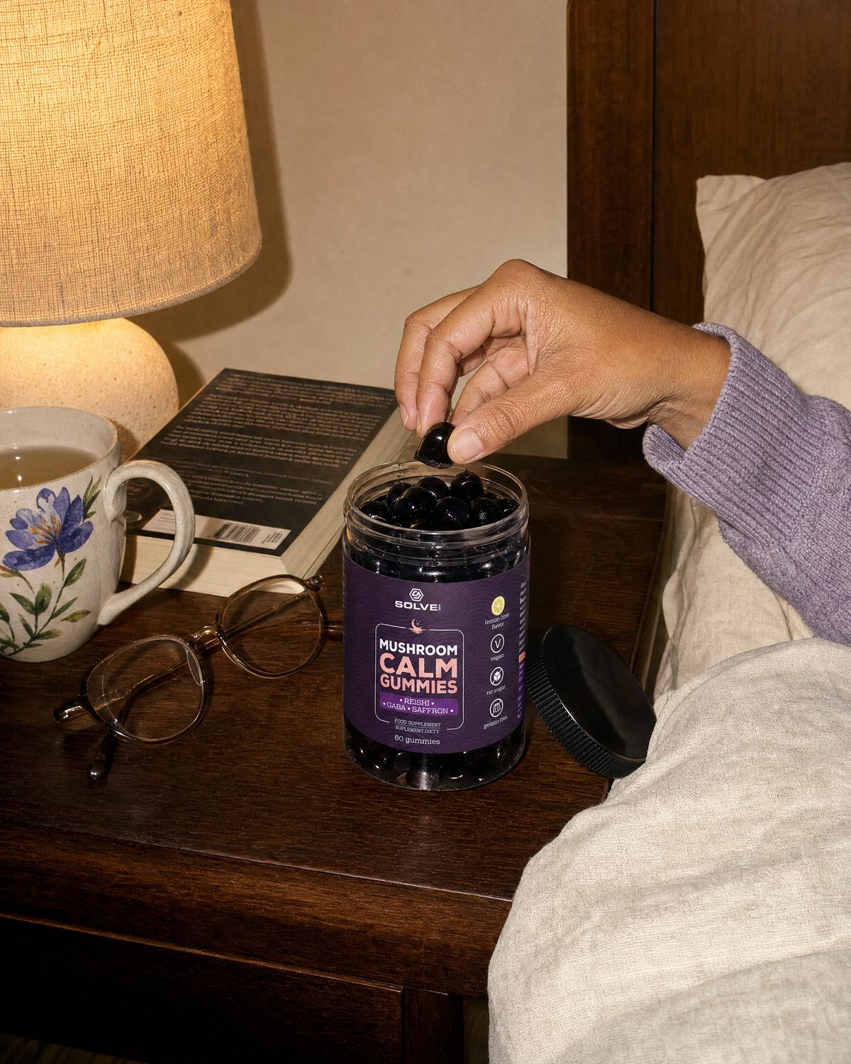 A hand lifting a gummy from the open jar on a bedside table beside a lamp, a book and a mug of tea