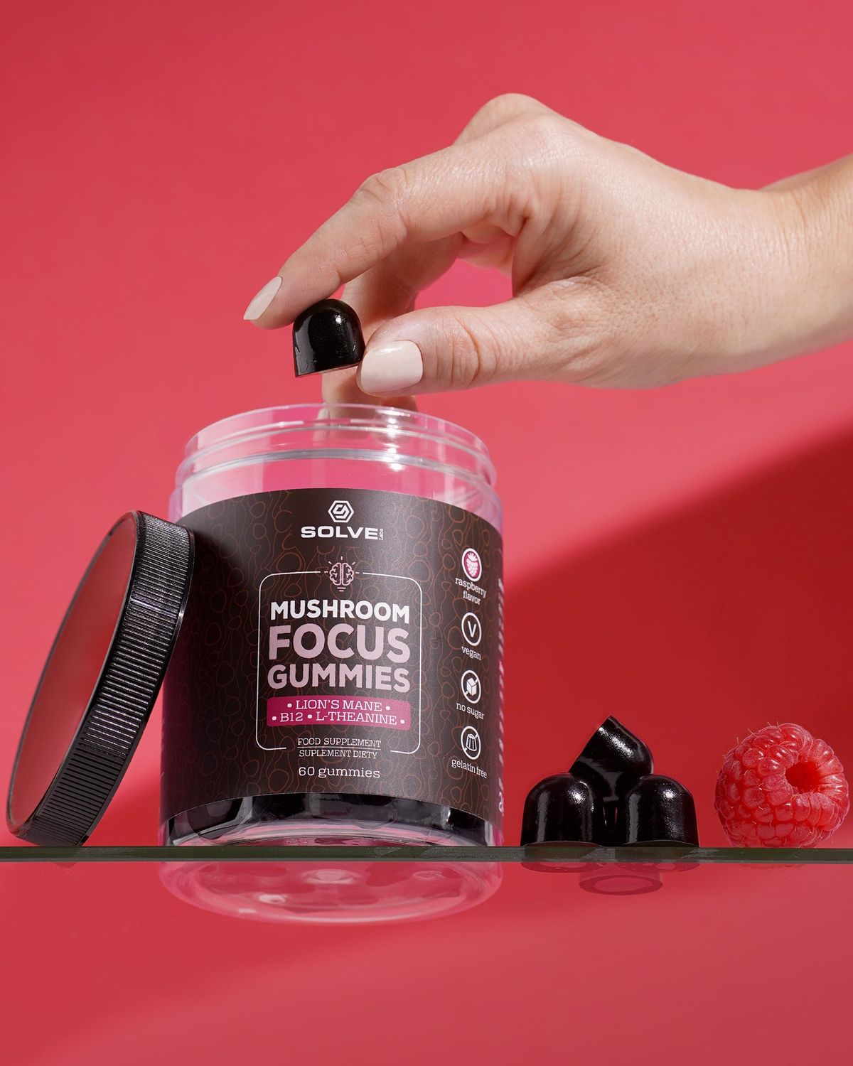 A hand lifting a gummy out of the open jar of Solve Labs Mushroom Focus Gummies on a red backdrop with raspberries