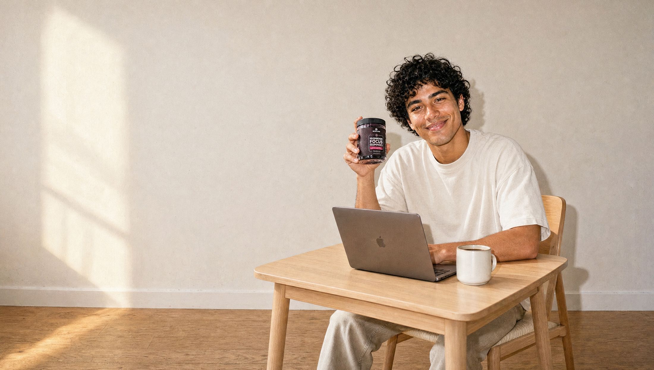 A young man at a sunlit desk with a laptop, holding up a gummy, the jar of Solve Labs Mushroom Focus Gummies beside him