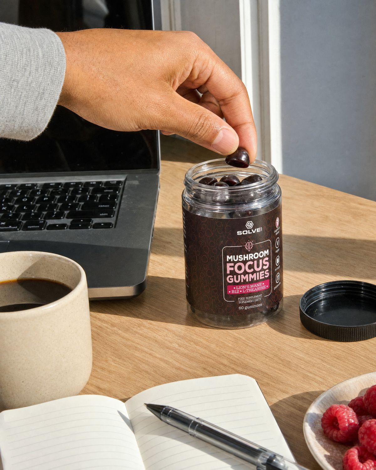 A hand lifting a gummy from the open jar on a desk beside a laptop and a notebook