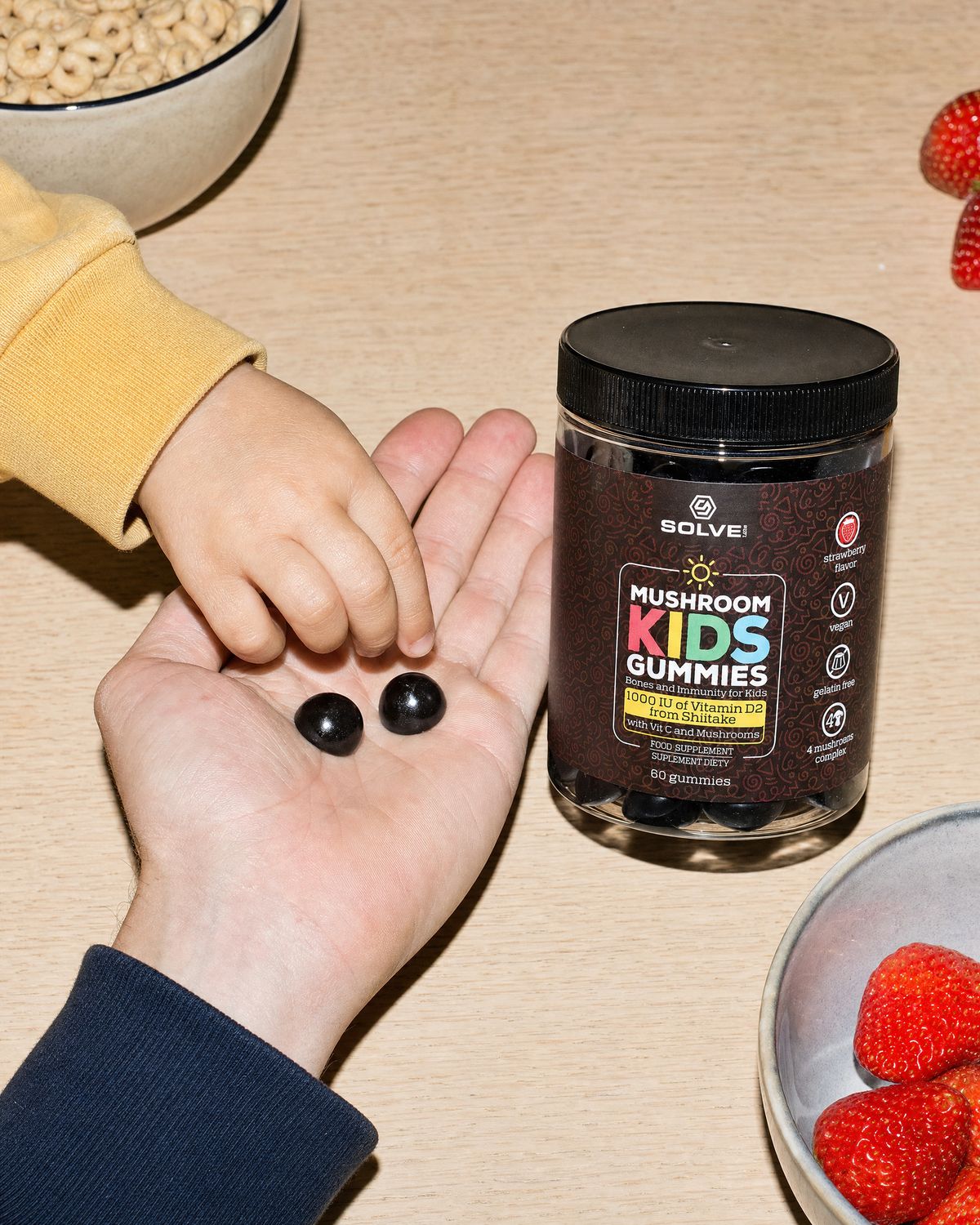A child taking a gummy from a parent&rsquo;s open palm at the breakfast table, the jar beside a glass of milk