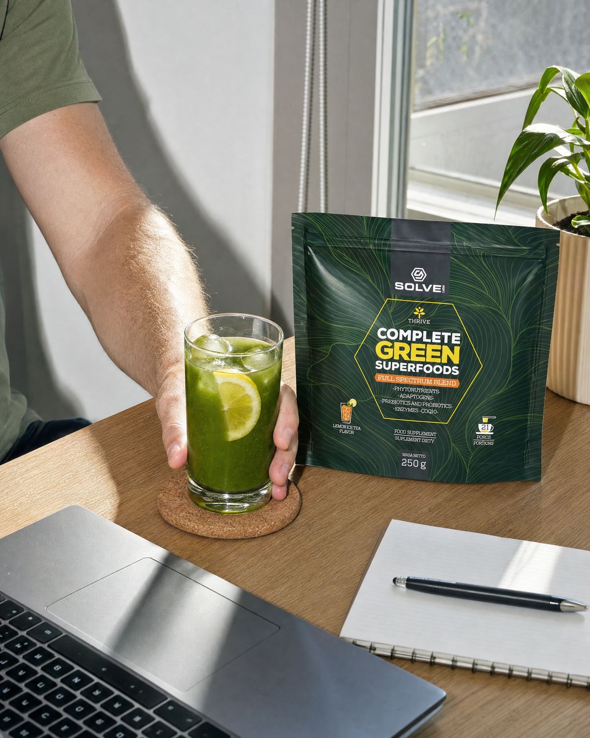 A hand setting a glass of green drink down on a desk beside a laptop and the pouch