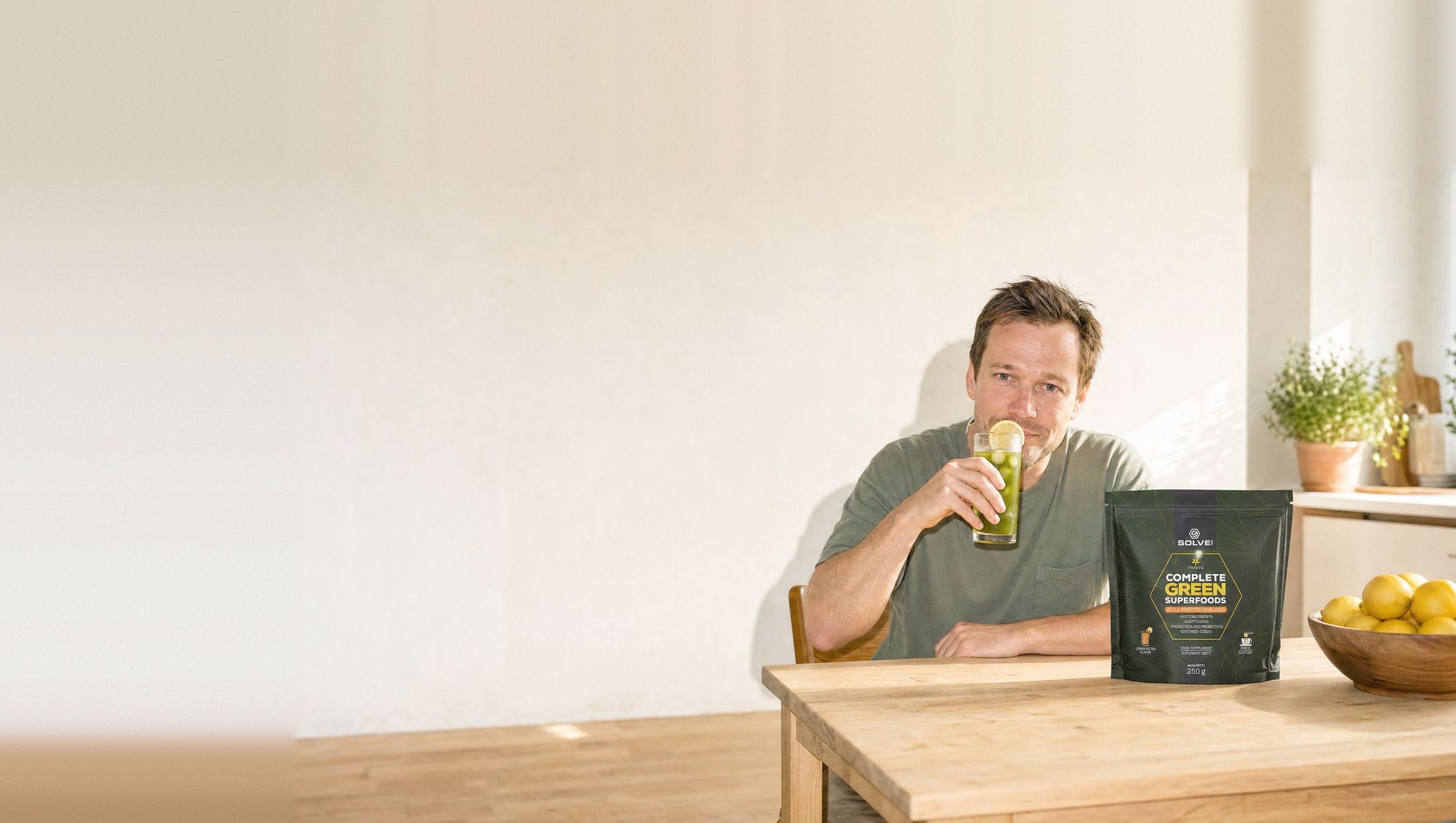 A man at a kitchen table in the morning drinking a tall glass of green drink, the pouch of Solve Labs Complete Green Superfoods beside him