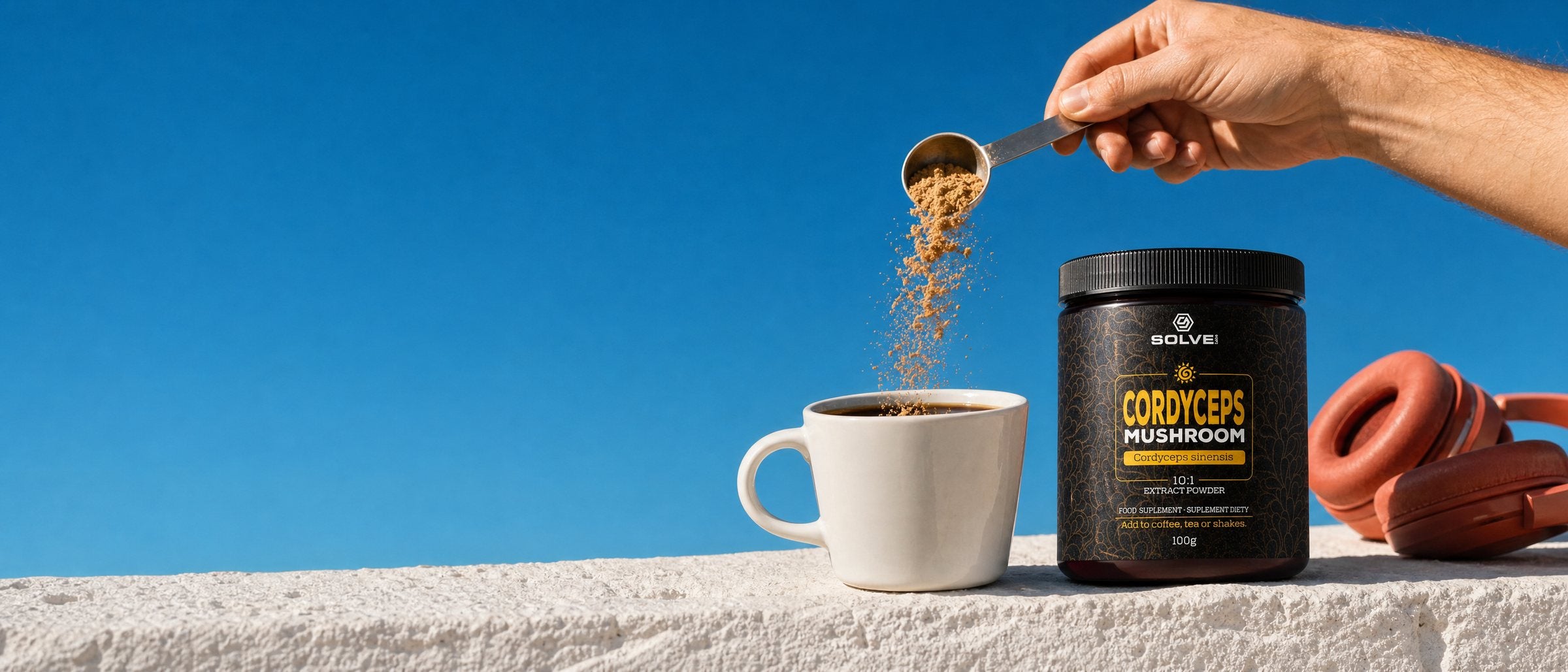A hand tipping a spoon of Cordyceps powder into a mug of coffee on a white wall under a blue sky, the jar of Solve Labs Cordyceps powder and a pair of headphones beside it