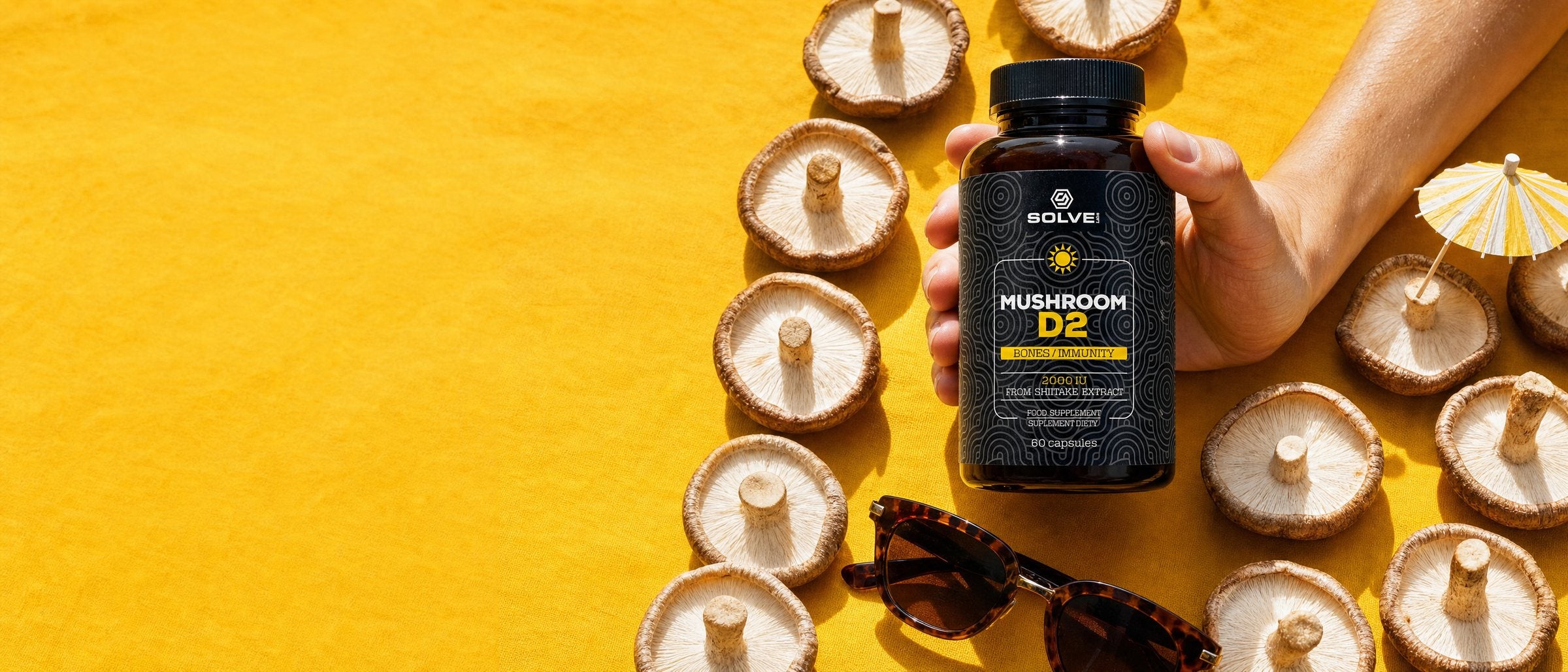 Shiitake mushrooms laid out in the sun on a yellow cloth with a pair of sunglasses and a paper umbrella, a hand standing the jar of Solve Labs Vitamin D2 among them