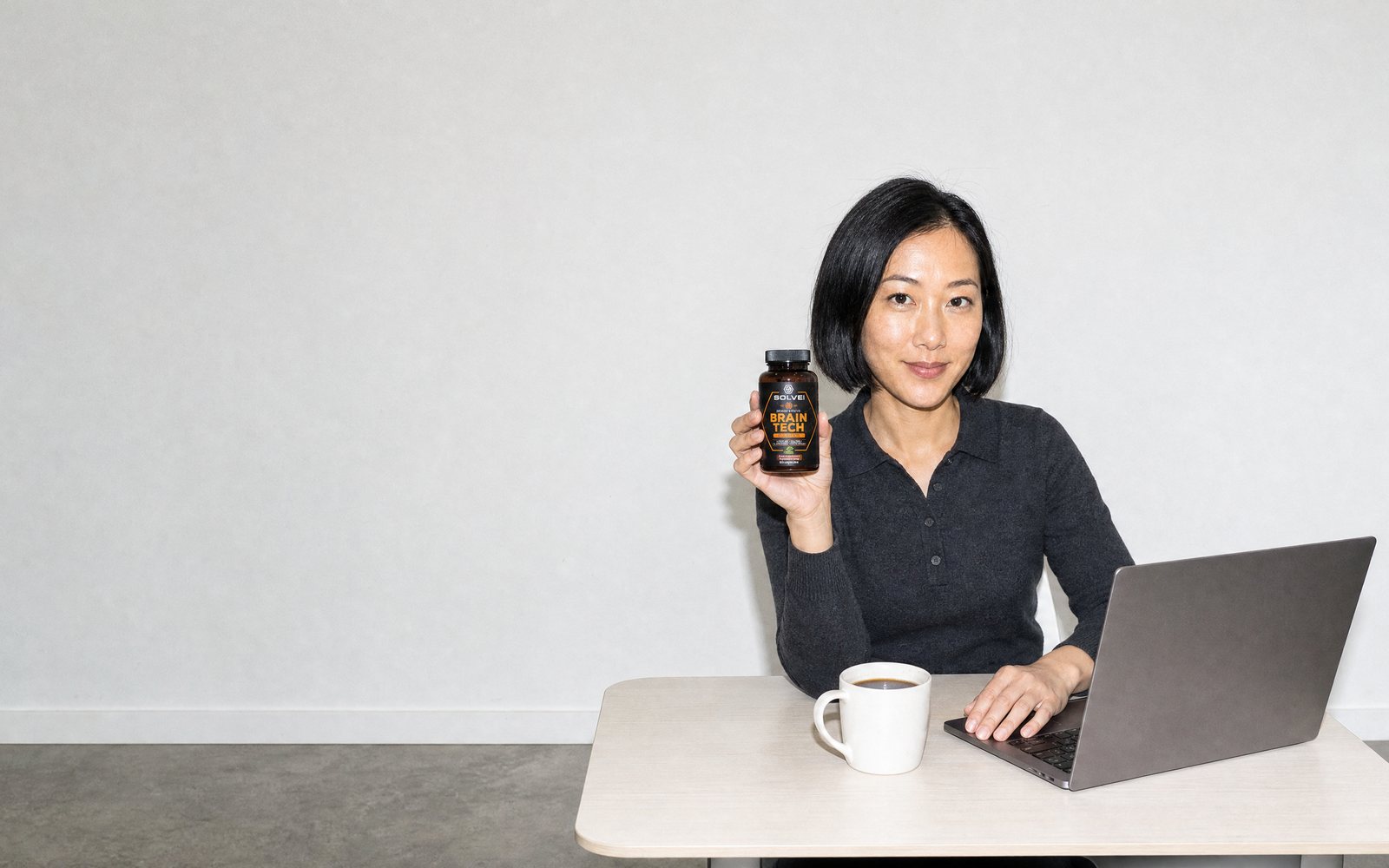 A woman at a desk in a bright office with a laptop and a cup of coffee, holding the Brain Tech jar