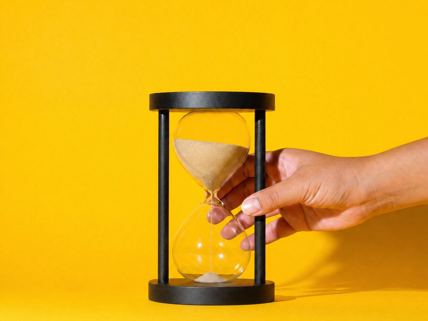 A hand setting down a sand hourglass on a yellow ground, the sand just starting to run