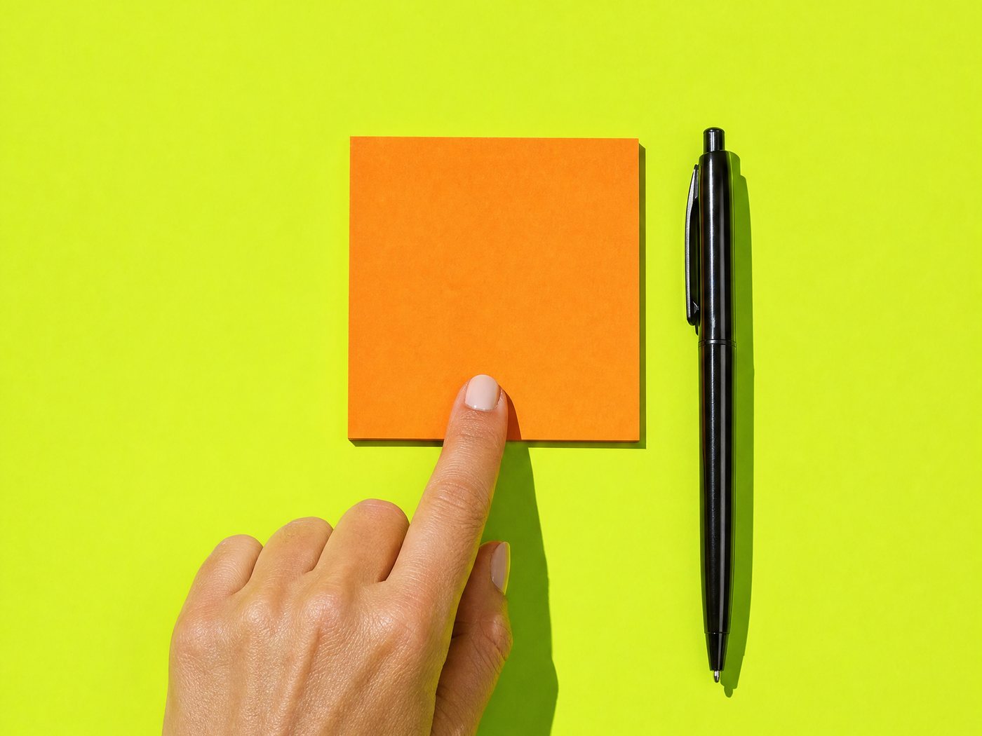 A fingertip pressing one blank orange sticky note onto a lime ground, a pen beside it