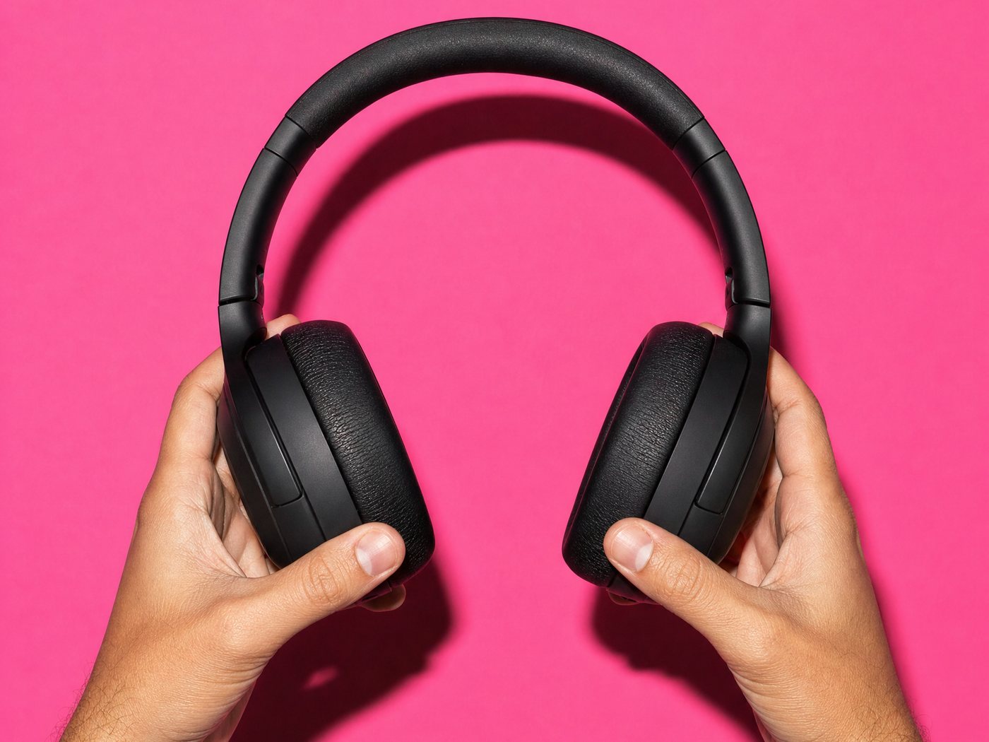 Two hands holding a pair of black over-ear headphones on a pink ground