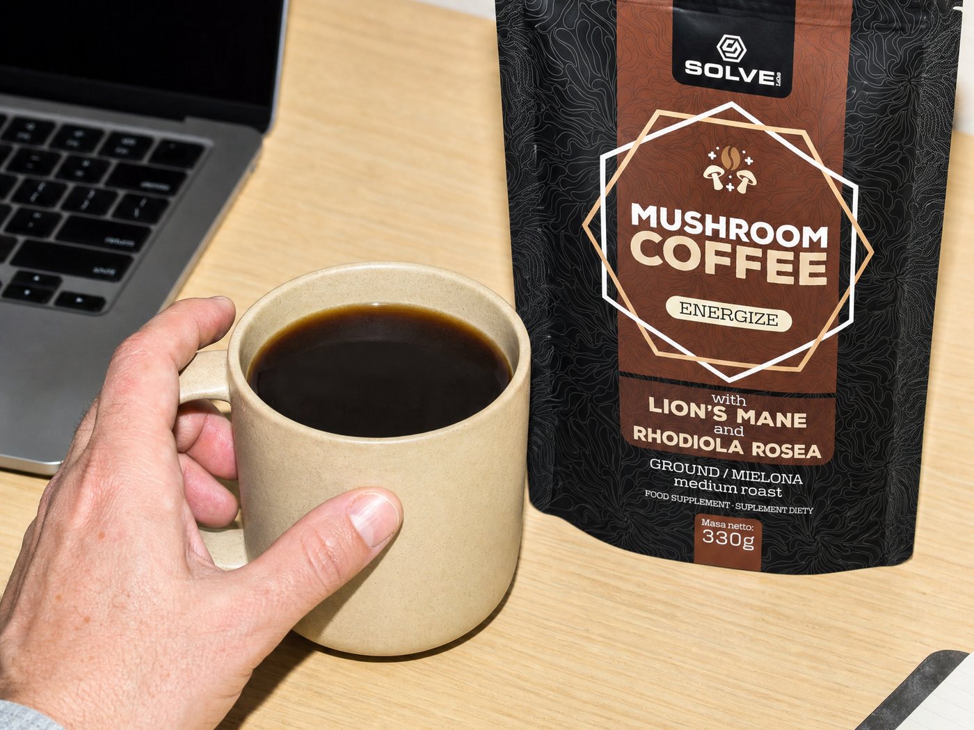 A hand lifting a mug of black coffee at a desk, the Mushroom Coffee bag beside a laptop