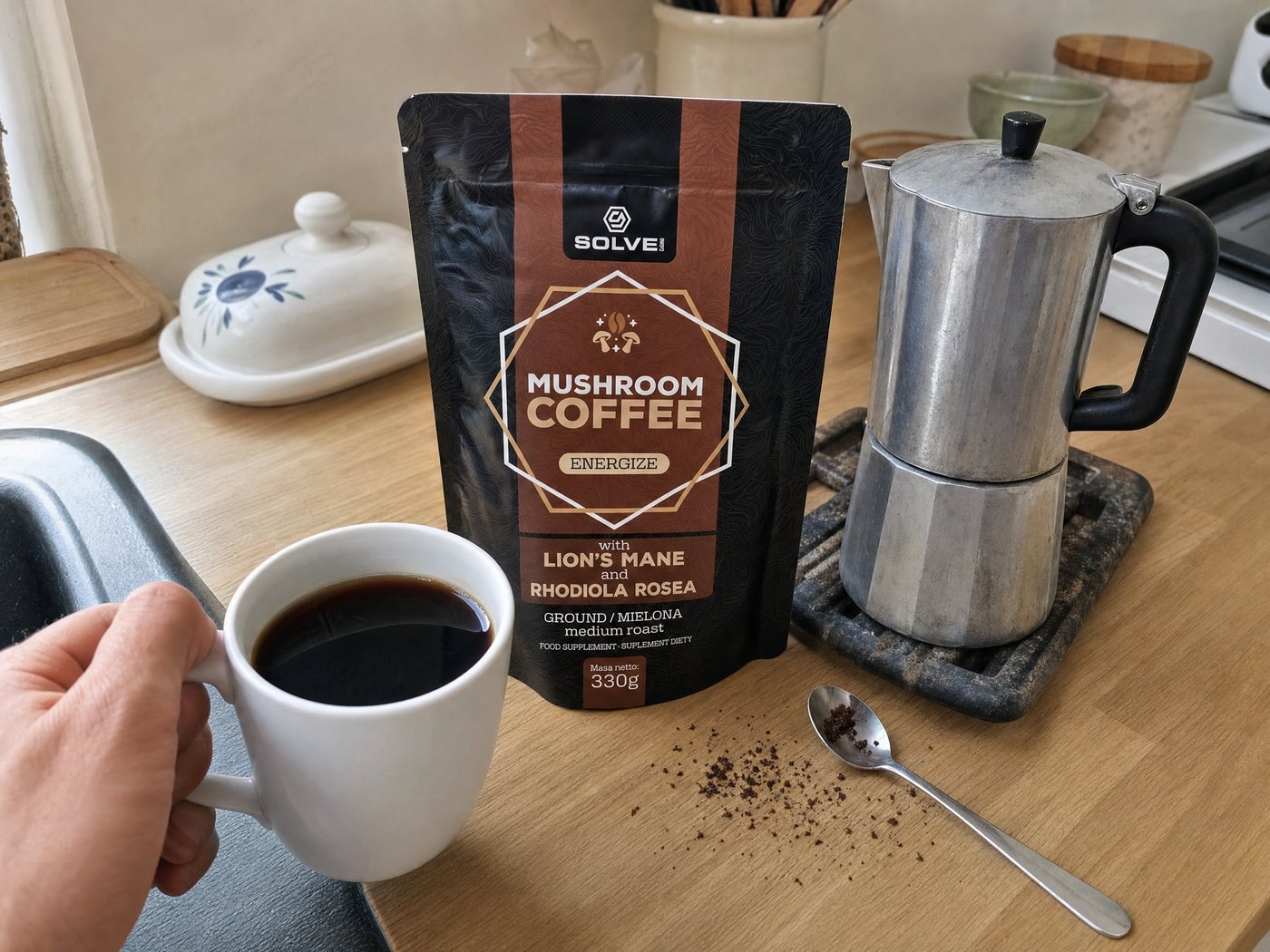 A hand lifting a mug of black coffee on a kitchen worktop beside the Mushroom Coffee bag and a moka pot