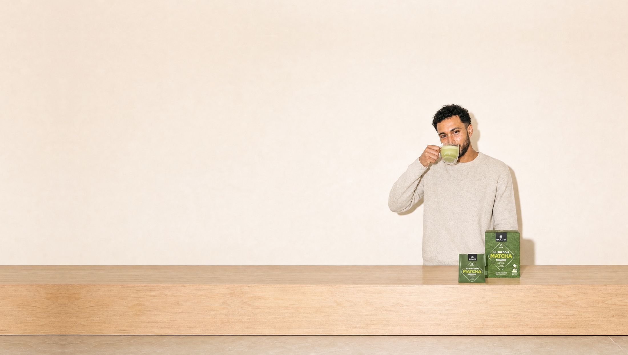 A man in a bright kitchen drinking a green matcha latte from a glass cup, the box of Solve Labs Mushroom Matcha and a sachet on the counter beside him