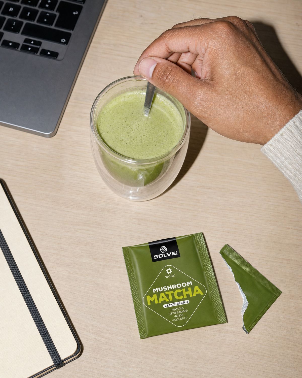 A hand stirring a green matcha latte in a glass cup on a desk beside a sachet and a laptop