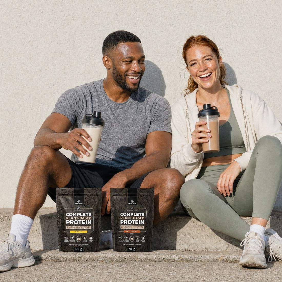 A man and a woman in gym clothes laughing on a bench with their shakers and two pouches of plant protein