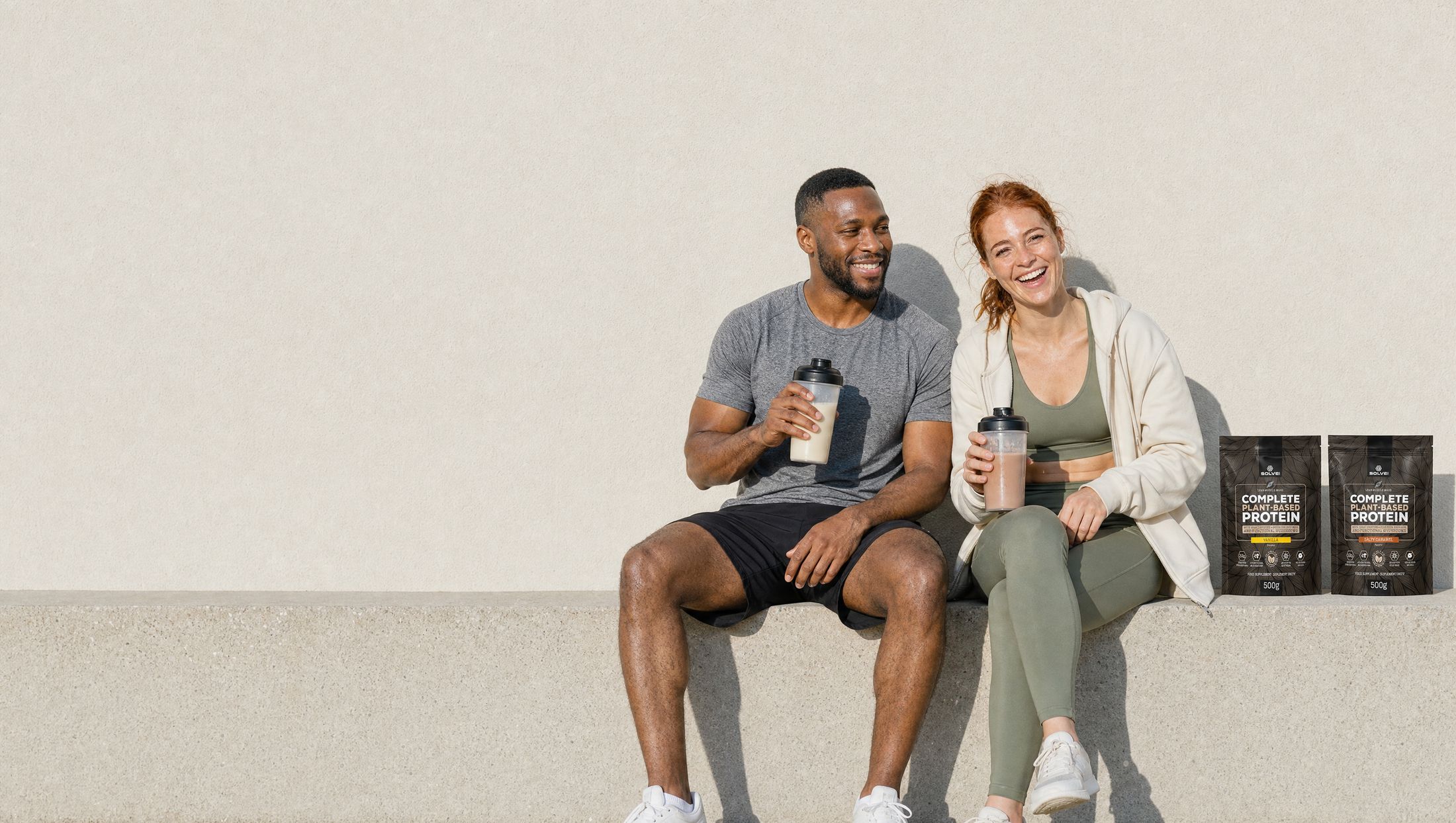 A man and a woman laughing on a sunlit step after a run, each with a shaker, the vanilla and salted caramel pouches of Solve Labs plant protein between them