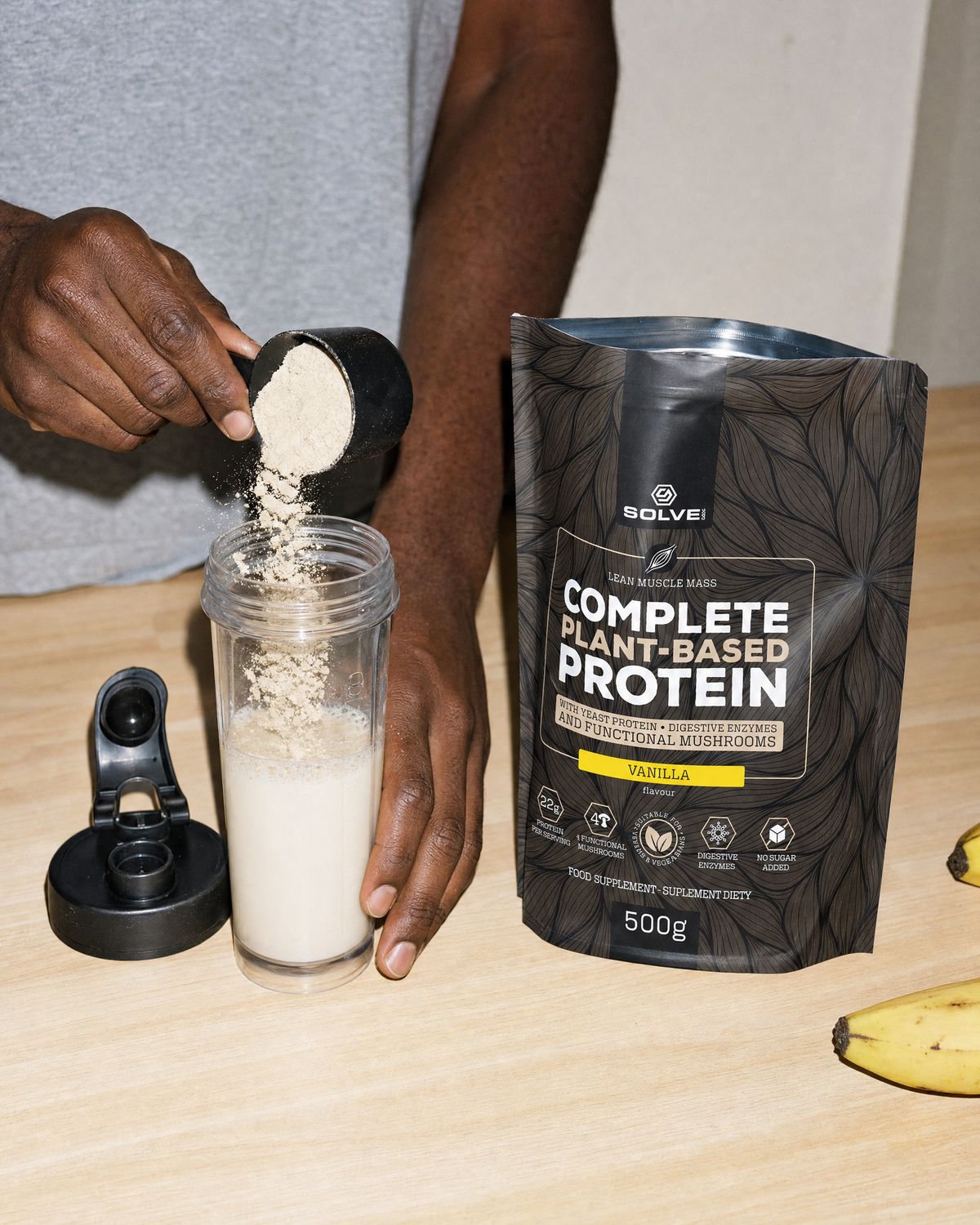 A scoop of vanilla protein powder falling into a shaker of oat milk beside the open pouch