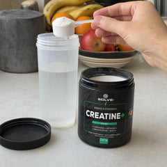 Une mesurette de Creatine+ tenue au-dessus d’un shaker d’eau, le pot ouvert à côté