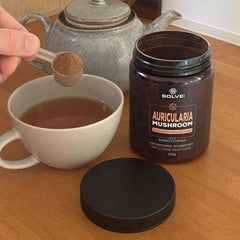 Une mesurette de poudre d’Auricularia tenue au-dessus d’une tasse de thé, le pot ouvert à côté