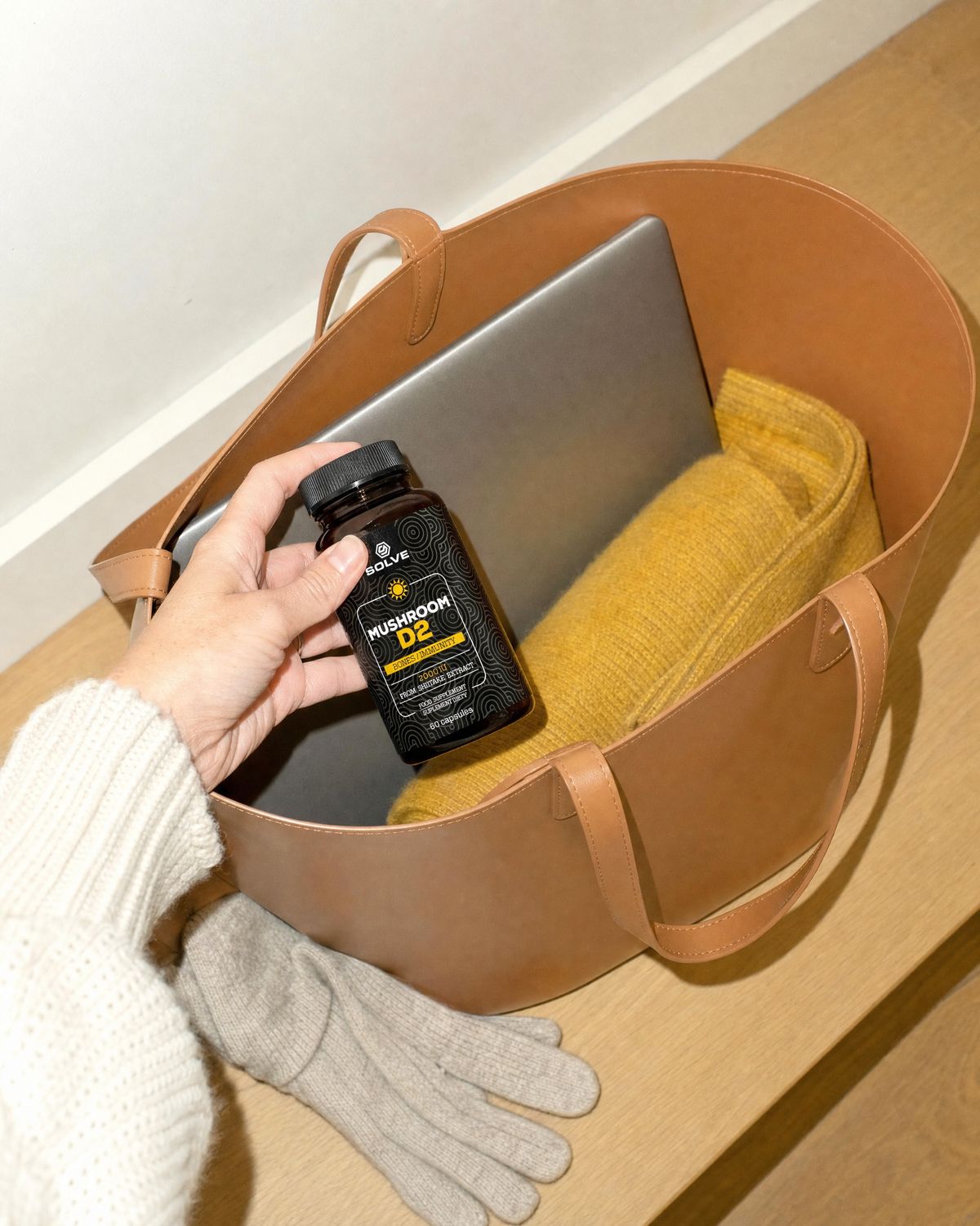 A hand dropping the jar into a leather tote bag beside a yellow scarf