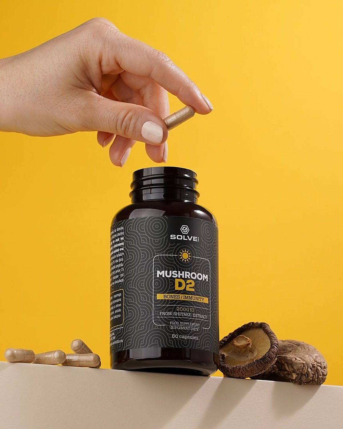 A hand dropping a capsule into the open jar of Solve Labs Mushroom D2 on a yellow backdrop, dried shiitake beside it
