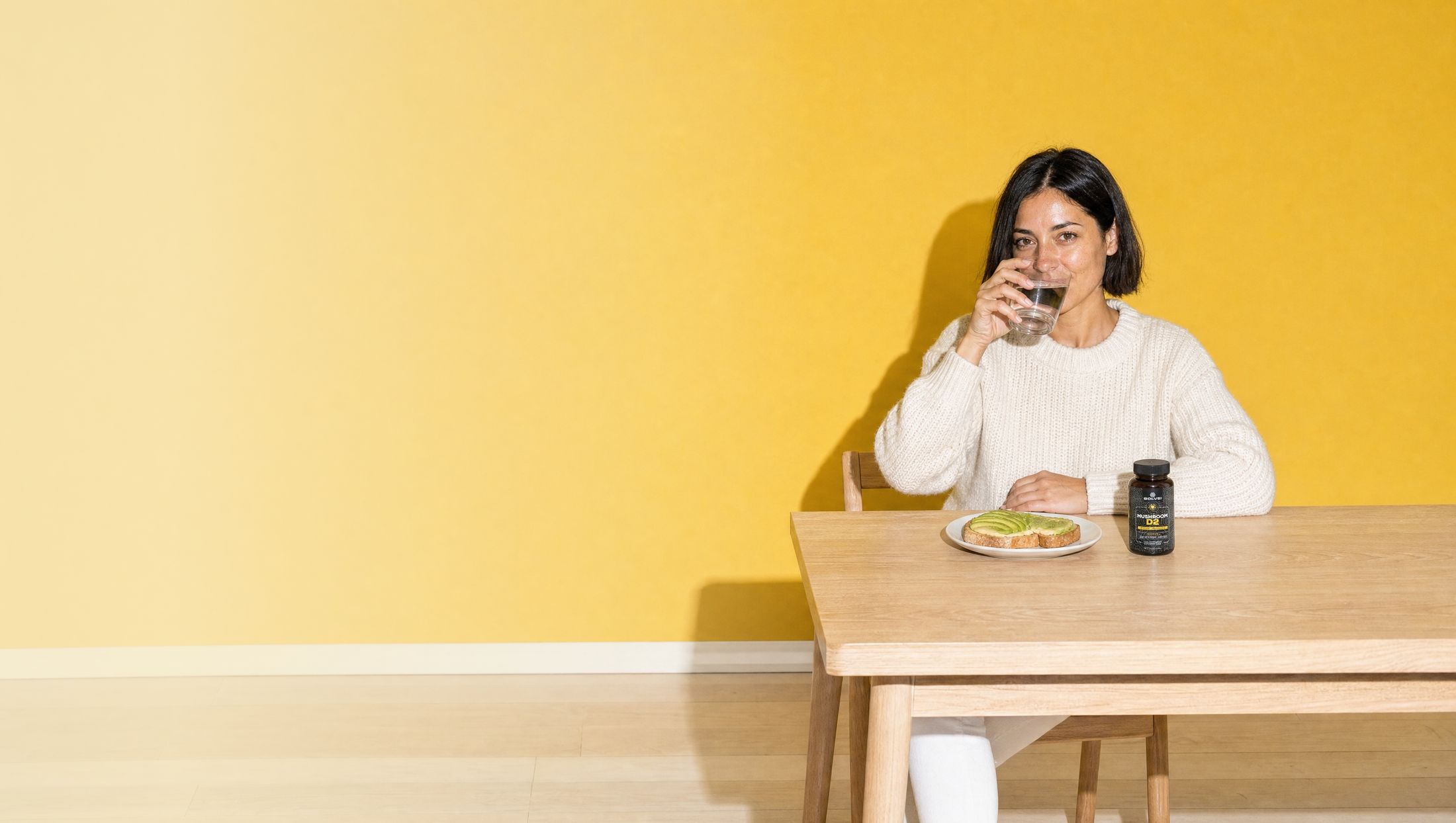 A woman at a breakfast table in front of a yellow wall with a glass of water, the jar of Solve Labs Mushroom D2 beside her plate