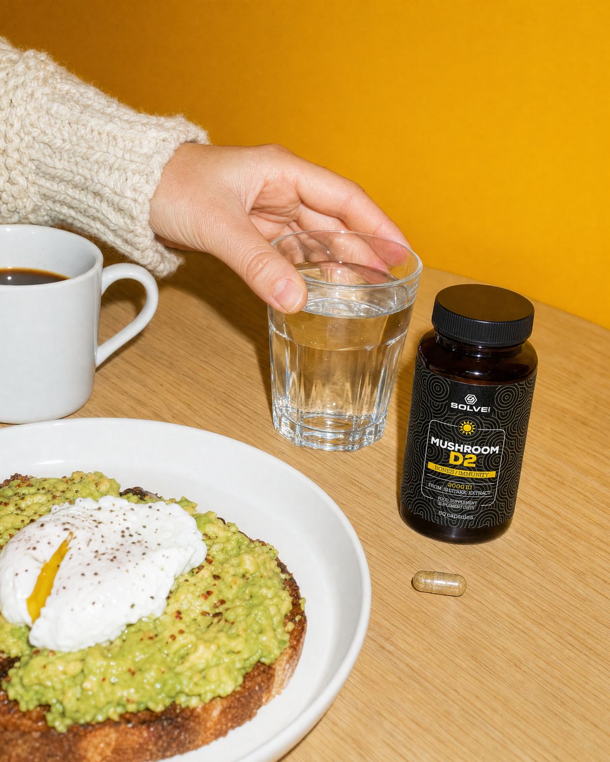 A hand reaching for a glass of water on a breakfast table beside the jar and a capsule