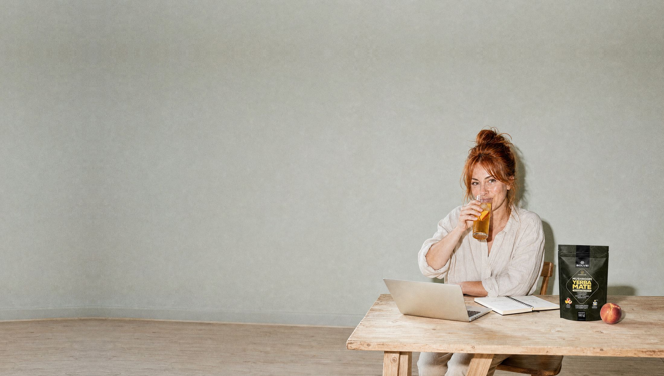 A woman at her desk in the afternoon drinking a tall glass of iced yerba mate, the pouch of Solve Labs Mushroom Yerba Mate beside her laptop