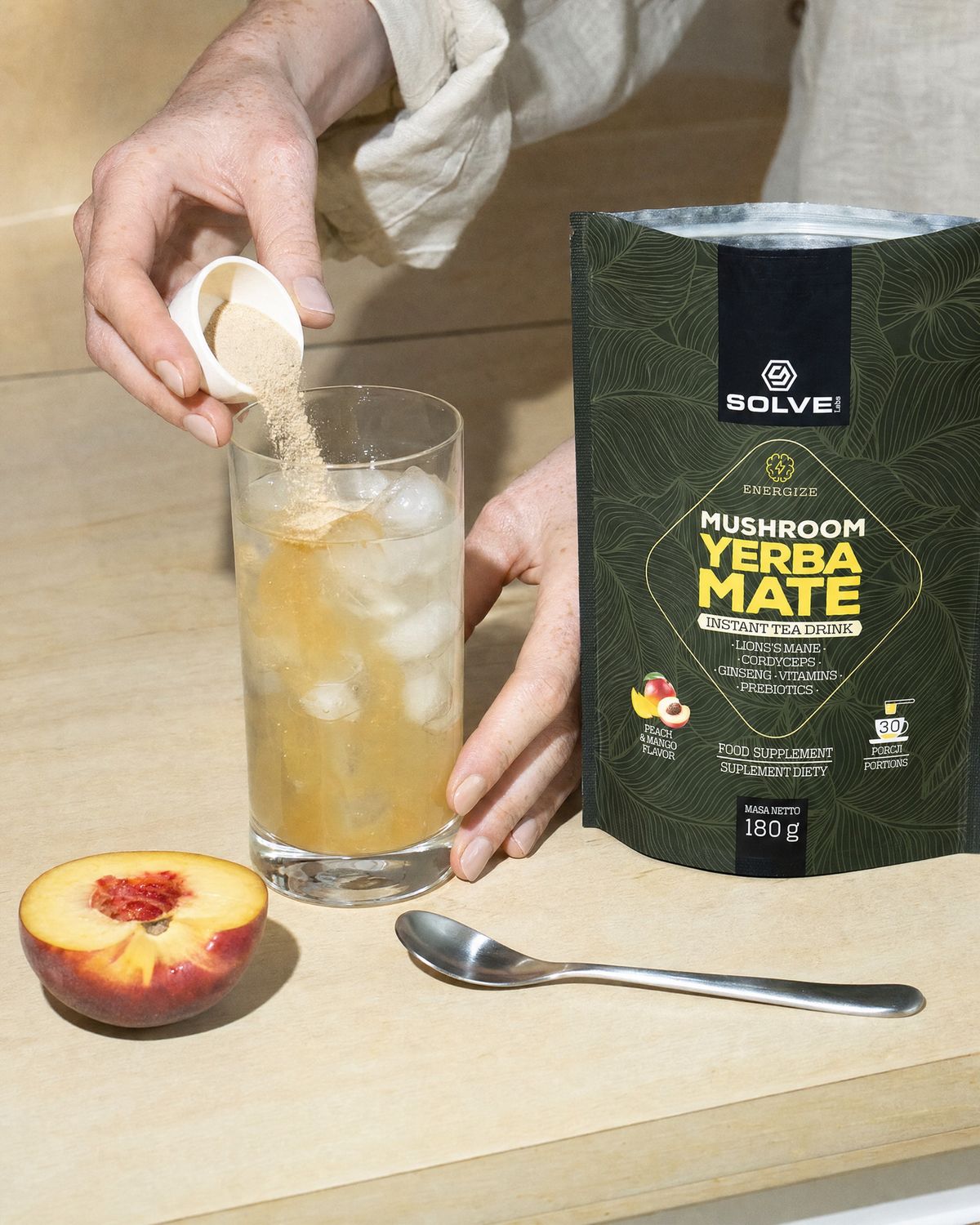 A scoop of pale powder falling into a tall glass of iced water beside the open pouch and a halved peach