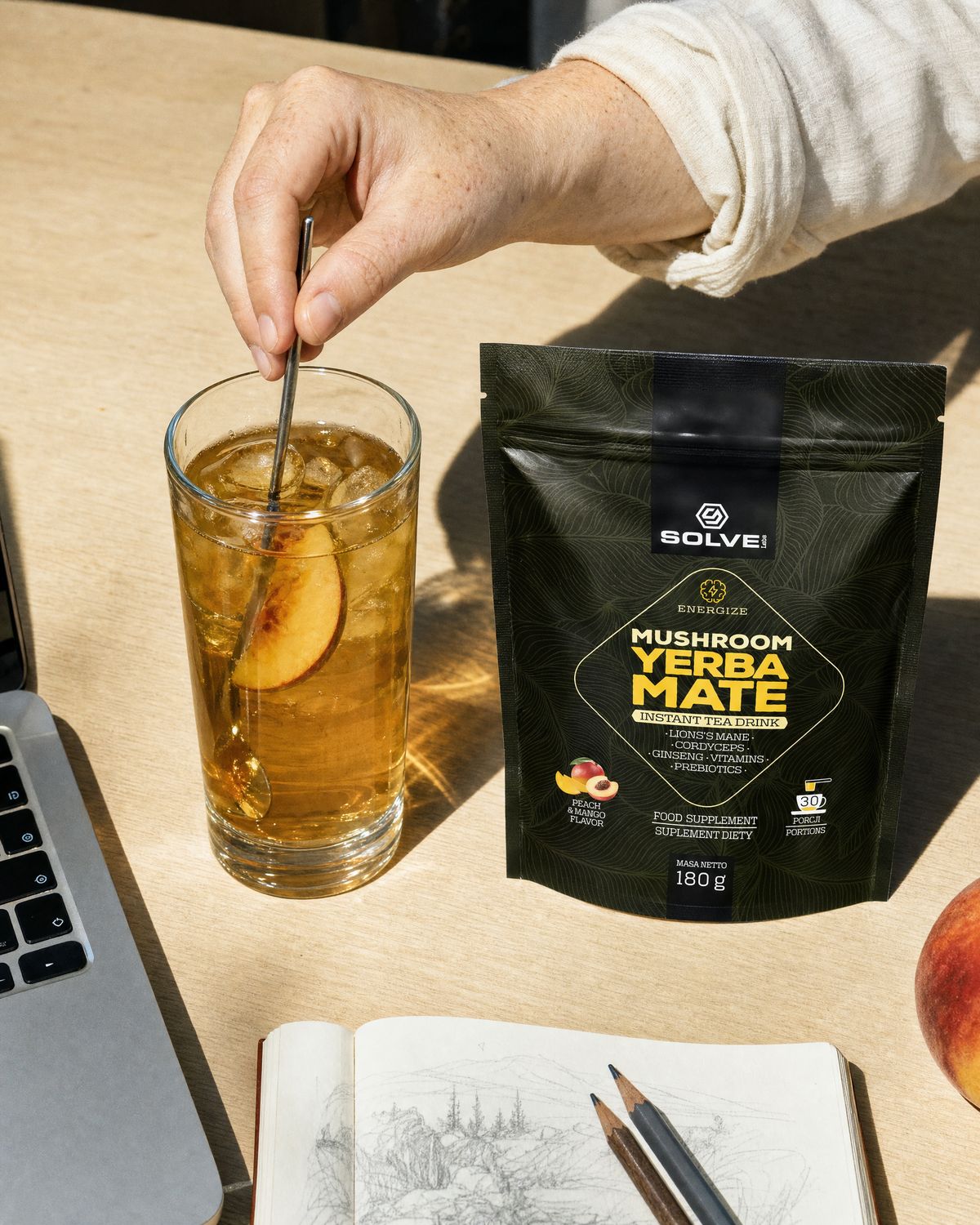 A hand stirring a tall glass of iced yerba mate on a desk beside the pouch